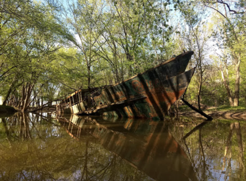 An abandoned, rusted shipwreck partially submerged in a calm river, surrounded by lush green trees.