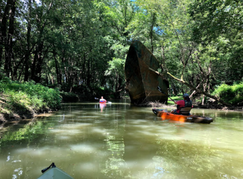 Two kayakers navigate a narrow, tree-lined waterway, passing a large, rusted shipwreck partially submerged in the water.