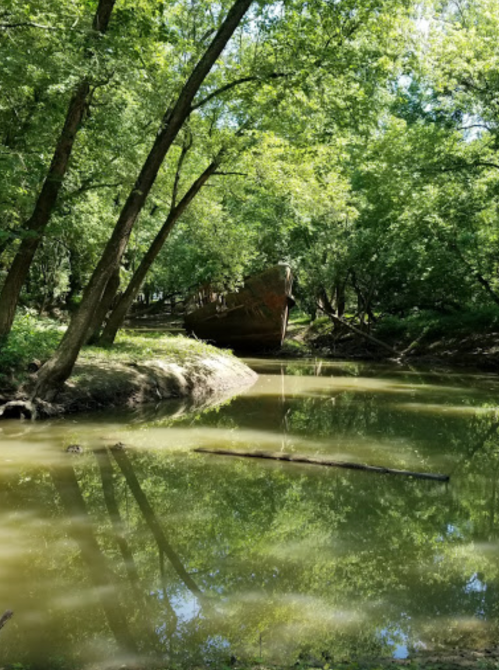 Abandoned ship partially submerged in a serene, green forested area with reflections in the calm water.