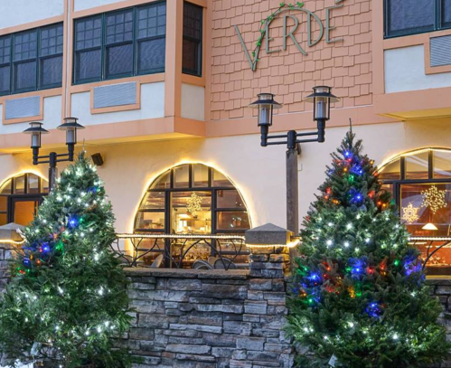 Two decorated Christmas trees with colorful lights stand outside a building with the sign "Verde" and warm lighting.