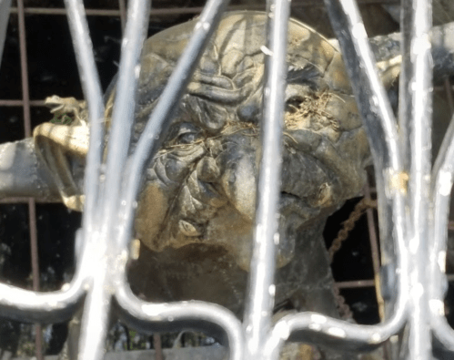 A weathered, grotesque face partially obscured by a decorative metal grate.