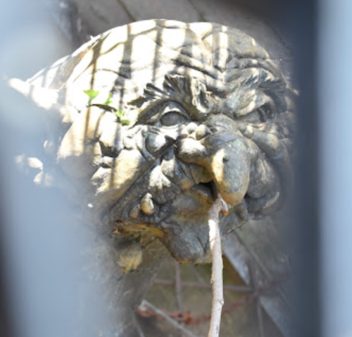 A close-up of a whimsical, weathered troll sculpture with a mischievous expression, partially obscured by bars.