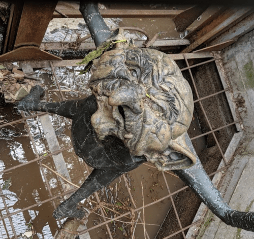 A weathered, grotesque sculpture resembling a face, partially submerged in muddy water and surrounded by debris.