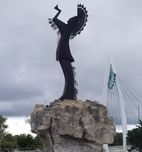 A tall black silhouette of a woman with a fan, standing on a rock against a cloudy sky.