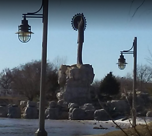 A tall statue on a rocky base, surrounded by trees and street lamps, near a body of water on a clear day.
