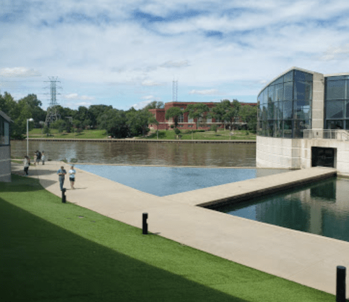 A serene riverside view featuring a modern building, green grass, and people walking along the path.