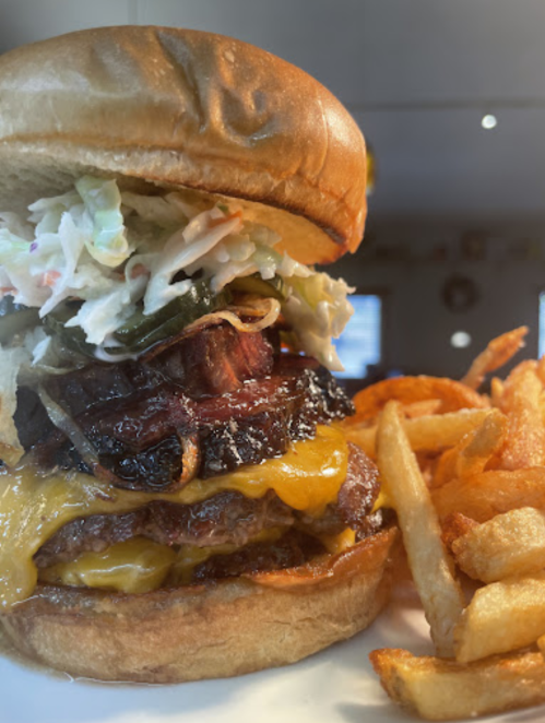 A towering burger with melted cheese, coleslaw, and grilled meat, served with crispy fries on the side.