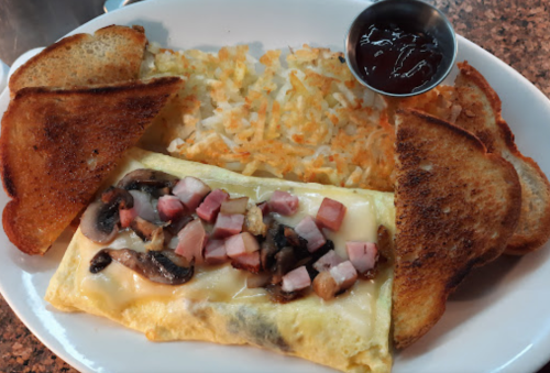 A plate featuring an omelette with ham and mushrooms, hash browns, and toast, served with a side of ketchup.