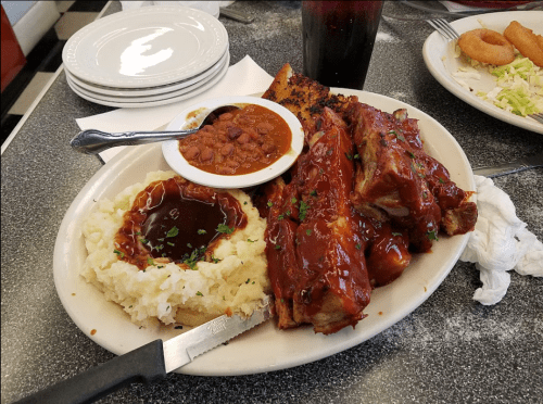 A plate of BBQ ribs with sauce, mashed potatoes, baked beans, and a drink, served on a diner table.