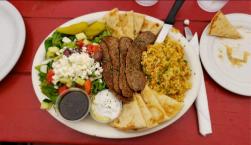 A plate of grilled meat slices, rice, salad, pita bread, pickles, and dipping sauces on a red table.