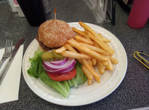 A sesame seed burger with lettuce, tomato, onion, pickles, and a side of golden French fries on a white plate.