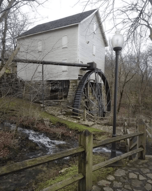 Historic watermill with a large wooden wheel, surrounded by trees and a small stream, near a wooden fence.