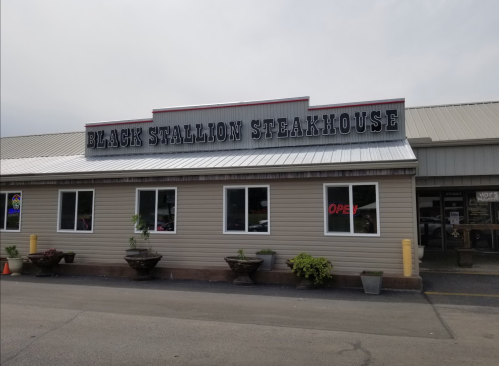 Exterior of Black Stallion Steakhouse with a sign and open neon light, featuring planters in front.