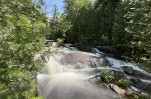 A serene waterfall cascades over rocks, surrounded by lush green trees under a clear blue sky.