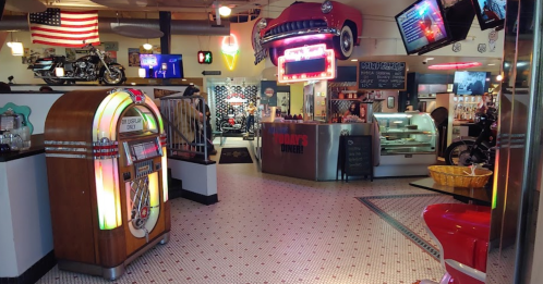 A retro diner interior featuring a jukebox, neon signs, and vintage decor, with motorcycles displayed in the background.