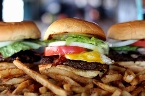 Three burgers with lettuce, tomato, onion, and a fried egg, served with a side of golden French fries.