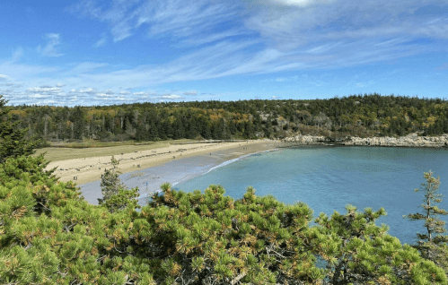 A panoramic view of a sandy beach surrounded by lush greenery and calm blue waters under a partly cloudy sky.