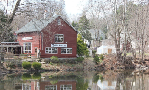 A red inn by a calm river, surrounded by trees and a few houses in the background on a clear day.