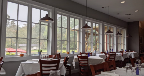 A cozy restaurant interior with white tablecloths, large windows, and pendant lights, overlooking a green landscape.