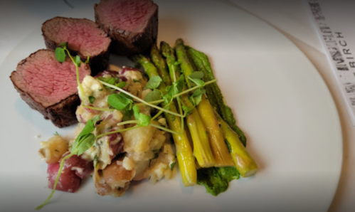 A plate featuring sliced beef tenderloin, asparagus, and a creamy potato salad garnished with microgreens.