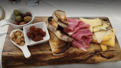 A wooden platter with toasted bread, cured meats, cheese slices, olives, and a small dish of red spread.