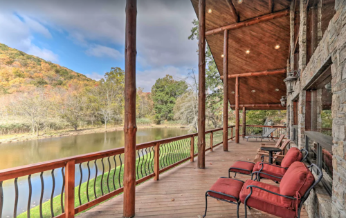 A wooden deck with red chairs overlooks a serene pond and lush hills under a partly cloudy sky.