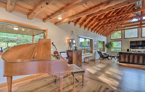 A cozy living space featuring a grand piano, wooden beams, large windows, and a comfortable seating area.