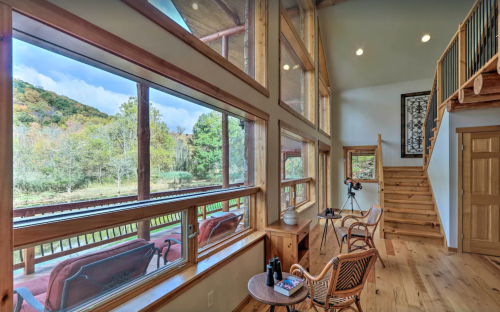 Cozy interior with large windows showcasing a scenic view of trees and hills, featuring a staircase and seating area.