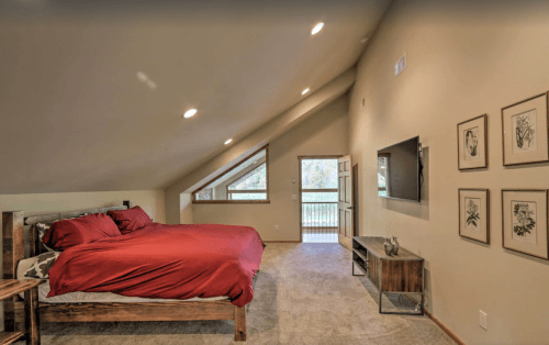 Cozy attic bedroom with a red bedspread, large windows, and a TV on the wall, featuring a warm, inviting atmosphere.