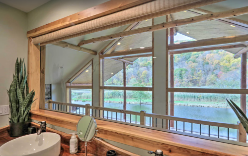 A bathroom mirror reflects a scenic view of a lake and colorful trees through large windows.