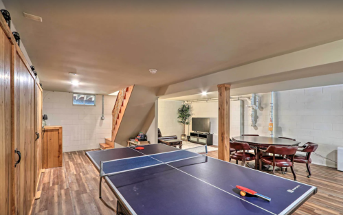 A spacious basement with a ping pong table, wooden accents, and a seating area with a TV in the background.