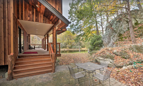 A wooden deck leads to a patio with a table and chairs, surrounded by autumn foliage and rocky terrain.