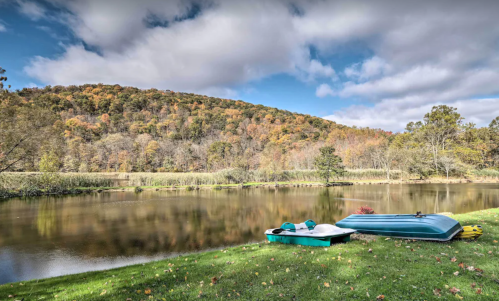 A serene lake surrounded by autumn foliage, with two boats resting on the grassy shore under a partly cloudy sky.