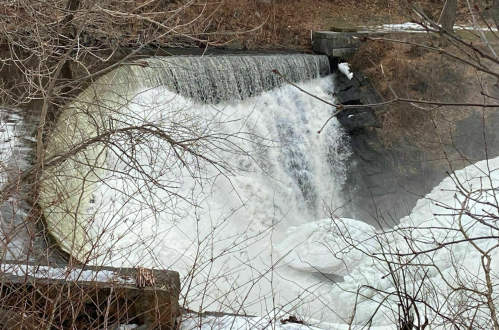 A frozen waterfall cascading over a dam, surrounded by bare trees and winter scenery.