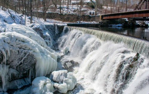 A frozen waterfall cascades over rocks, surrounded by snow-covered trees and a bridge in the background.
