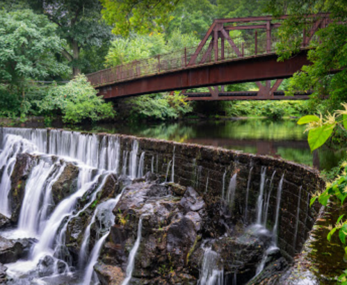 A serene waterfall cascades over rocks, with a rustic bridge arching over a calm river surrounded by lush greenery.