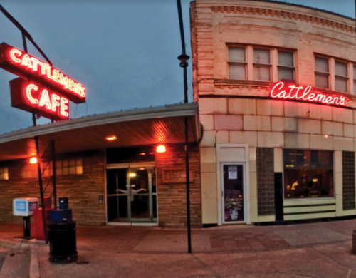 A cafe with neon signs reading "Cattlemen's Cafe" and "Cattlemen's," illuminated at dusk.