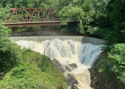 A waterfall cascades over rocks beneath a rusted train bridge, surrounded by lush green trees.