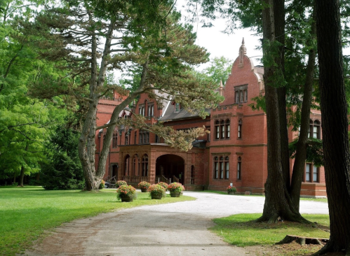 A large red brick mansion surrounded by lush green trees and flower planters along a gravel path.