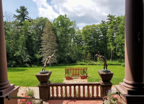 View from a porch featuring two statues and a lush green garden surrounded by trees under a partly cloudy sky.