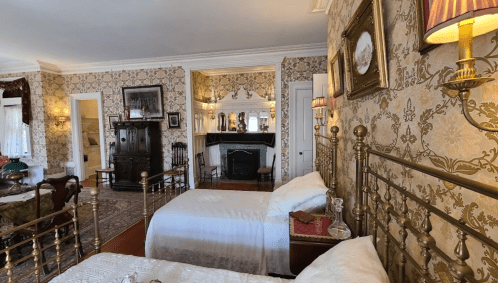 A vintage bedroom with two brass beds, ornate wallpaper, and a fireplace, featuring classic decor and elegant furnishings.