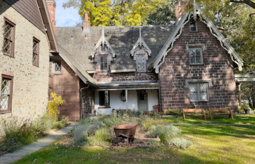 Historic stone house with a steep roof, decorative gables, and a garden featuring a wheelbarrow in the foreground.