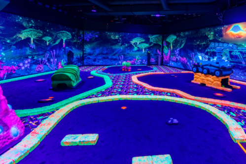 A vibrant glow-in-the-dark mini-golf course with colorful obstacles and a cosmic-themed backdrop.