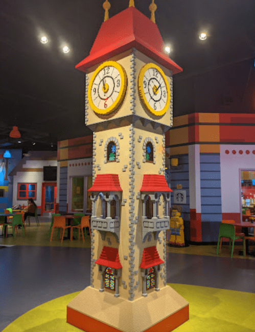 A colorful LEGO clock tower with two large clocks, surrounded by a playful, vibrant indoor space.