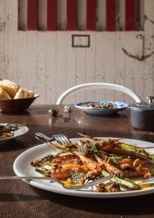 A plate of vibrant roasted carrots and asparagus on a rustic wooden table, with side dishes and bread in the background.