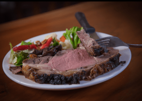 A plate with a slice of roast beef, accompanied by a fresh salad and vegetables on a wooden table.