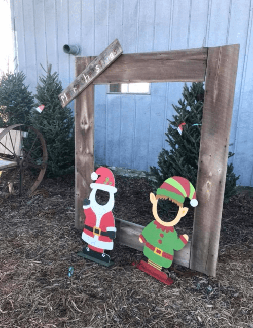 A wooden frame with cutouts of Santa and an elf, surrounded by Christmas trees and festive decorations.