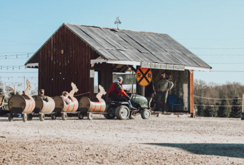 A rustic shed with a railroad sign, a person on a tractor, and barrels lined up outside in a sunny setting.