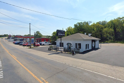 A small donut shop with a sign, surrounded by trees and parked cars along a road.