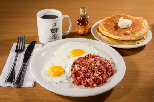A plate with two fried eggs, corned beef hash, pancakes with butter, and a cup of coffee on a wooden table.